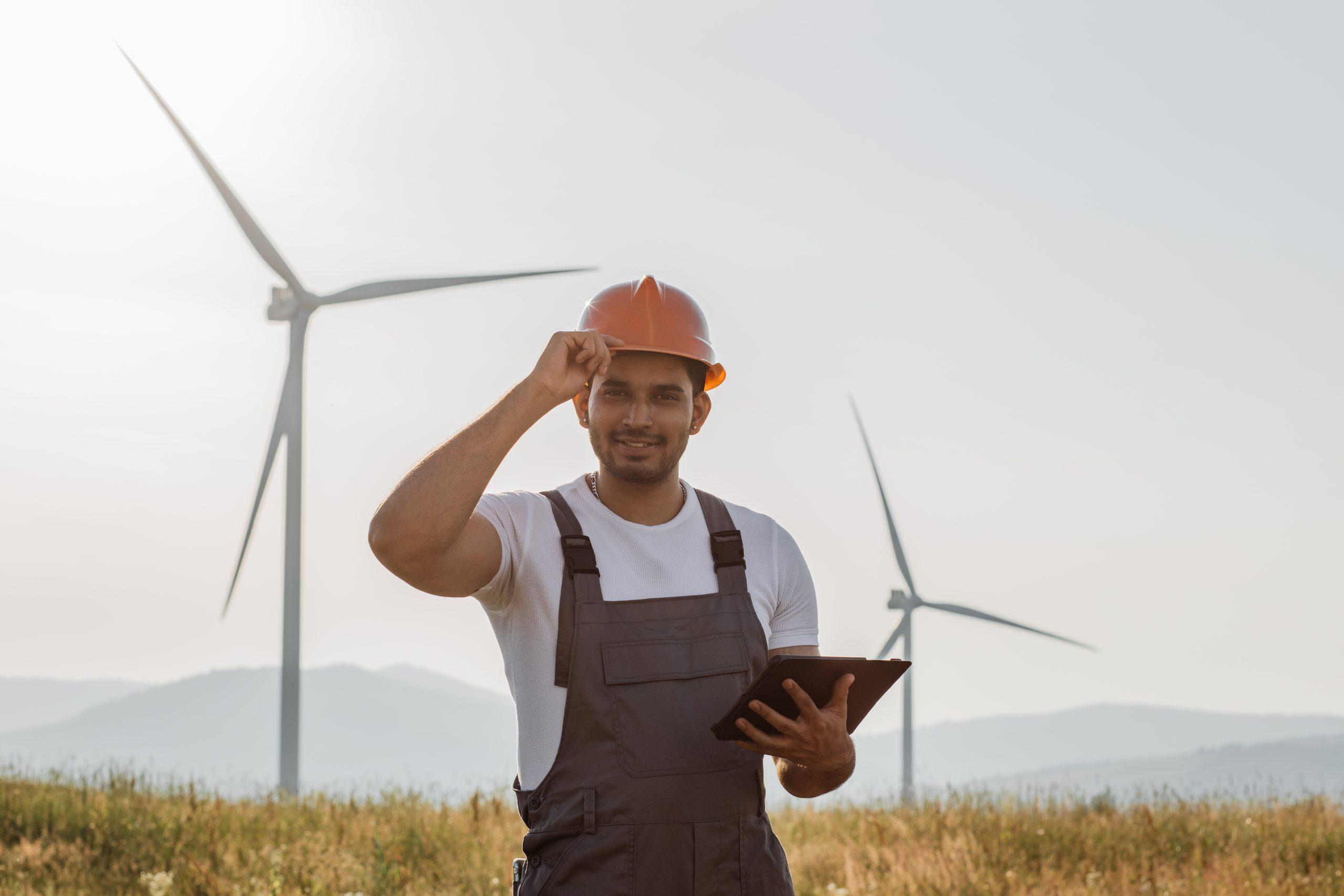 Professional technician in helmet and overalls smiling oj camera while standing on eco farm with wind turbines. Indian man using digital tablet while controlling process of clean energy making.